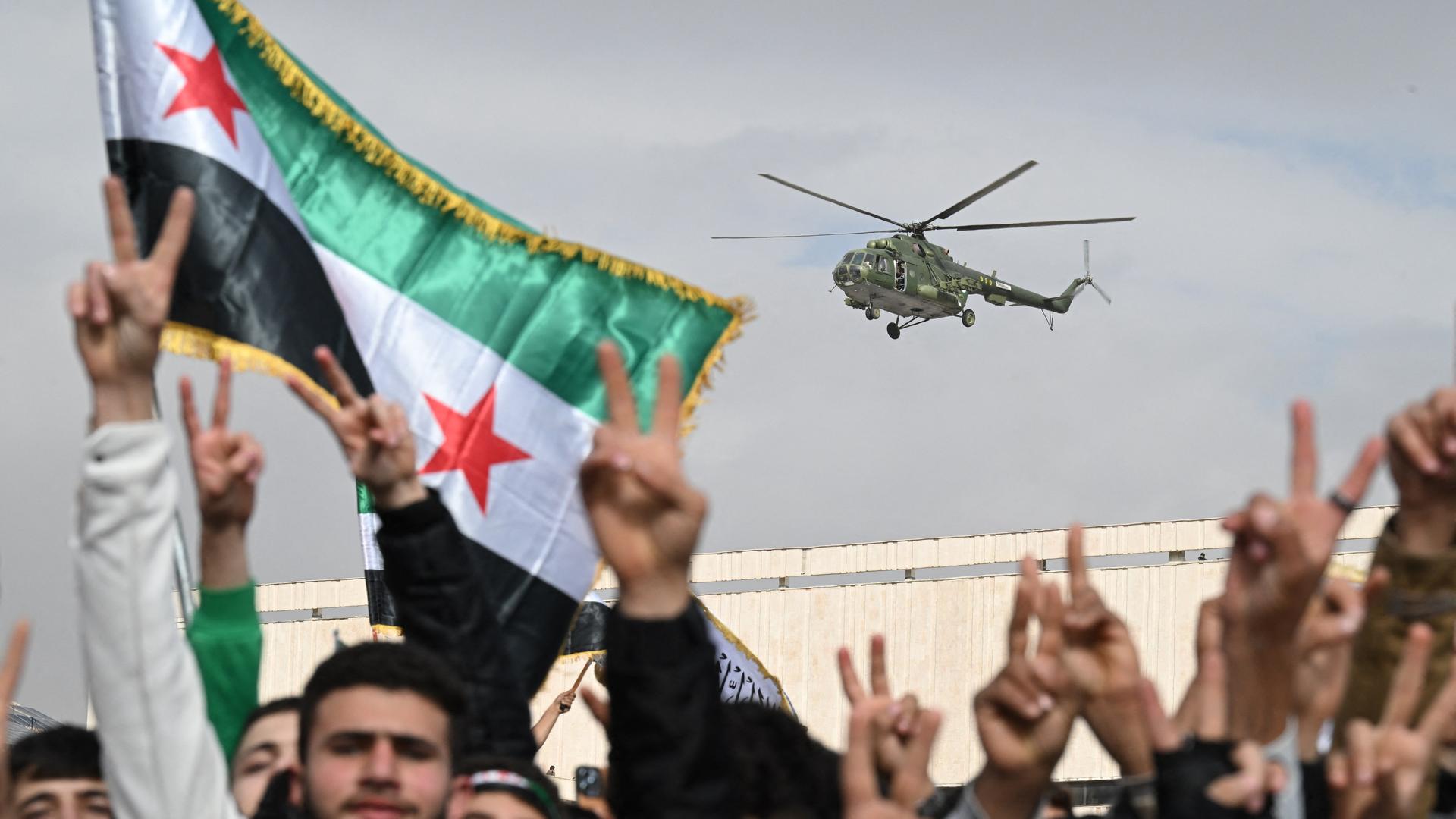 Ein Hubschrauber fliegt über eine Menschenmenge. Das Siegeszeichen und Flaggen werden gezeigt.  Es handelt sich um Feierlichkeiten zum Jahrestag des Sturzes des Machthabers Baschar al-Assad in der syrischen Hauptstadt Damaskus (8.12.2025, Syrien)