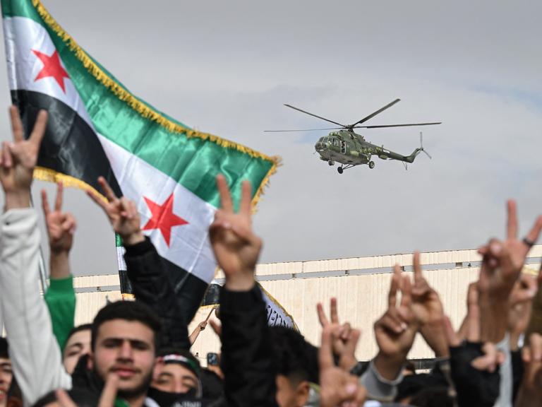 Ein Hubschrauber fliegt über eine Menschenmenge. Das Siegeszeichen und Flaggen werden gezeigt. Es handelt sich um Feierlichkeiten zum Jahrestag des Sturzes des Machthabers Baschar al-Assad in der syrischen Hauptstadt Damaskus (8.12.2025, Syrien) Ein Hubschrauber fliegt über eine Menschenmenge. Das Siegeszeichen und Flaggen werden gezeigt. Es handelt sich um Feierlichkeiten zum Jahrestag des Sturzes des Machthabers Baschar al-Assad in der syrischen Hauptstadt Damaskus (8.12.2025, Syrien)