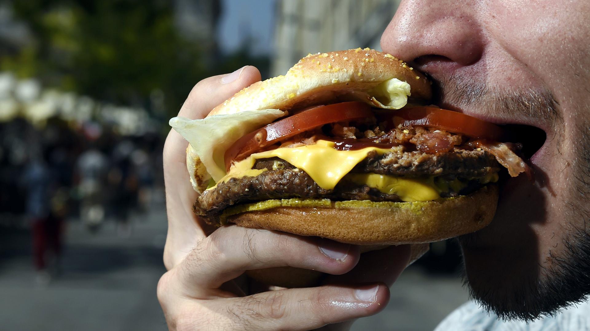 Ein bärtiger Mann beißt in einen Hamburger. Sein Mund ist weit geöffnet, der Hamburger ist üppig belegt. Eine Käsescheibe, eine Tomatenscheibe und Salat sind zu sehen. Auch mehrere Frikadellen sind zwischen den Brötchenhälften.