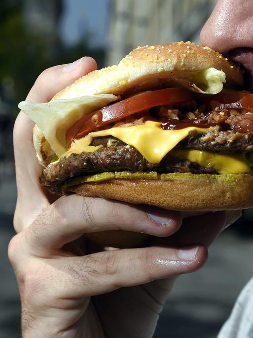 Ein bärtiger Mann beißt in einen Hamburger. Sein Mund ist weit geöffnet, der Hamburger ist üppig belegt. Eine Käsescheibe, eine Tomatenscheibe und Salat sind zu sehen. Auch mehrere Frikadellen sind zwischen den Brötchenhälften.