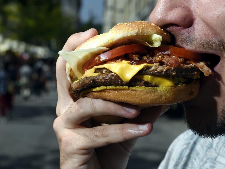 Ein bärtiger Mann beißt in einen Hamburger. Sein Mund ist weit geöffnet, der Hamburger ist üppig belegt. Eine Käsescheibe, eine Tomatenscheibe und Salat sind zu sehen. Auch mehrere Frikadellen sind zwischen den Brötchenhälften.