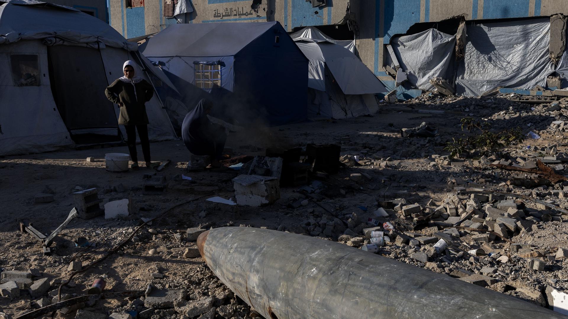 Im Vordergrund liegt eine Bombe, die von der israelischen Armee zurückgelassen wurde und nicht explodiert ist. Sie zeigt im Hintergrund auf Flüchtlingszelte im Gazastreife. Davor steht eine Person. Im Vordergrund liegt eine Bombe, die von der israelischen Armee zurückgelassen wurde und nicht explodiert ist. Sie zeigt im Hintergrund auf Flüchtlingszelte im Gazastreife. Davor steht eine Person.