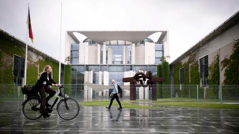 Außenansicht des Bundeskanzleramtes, es wirkt regnerisch. Ein Mann spaziert vorbei und eine Frau fährt auf dem Fahrrad vorbei.