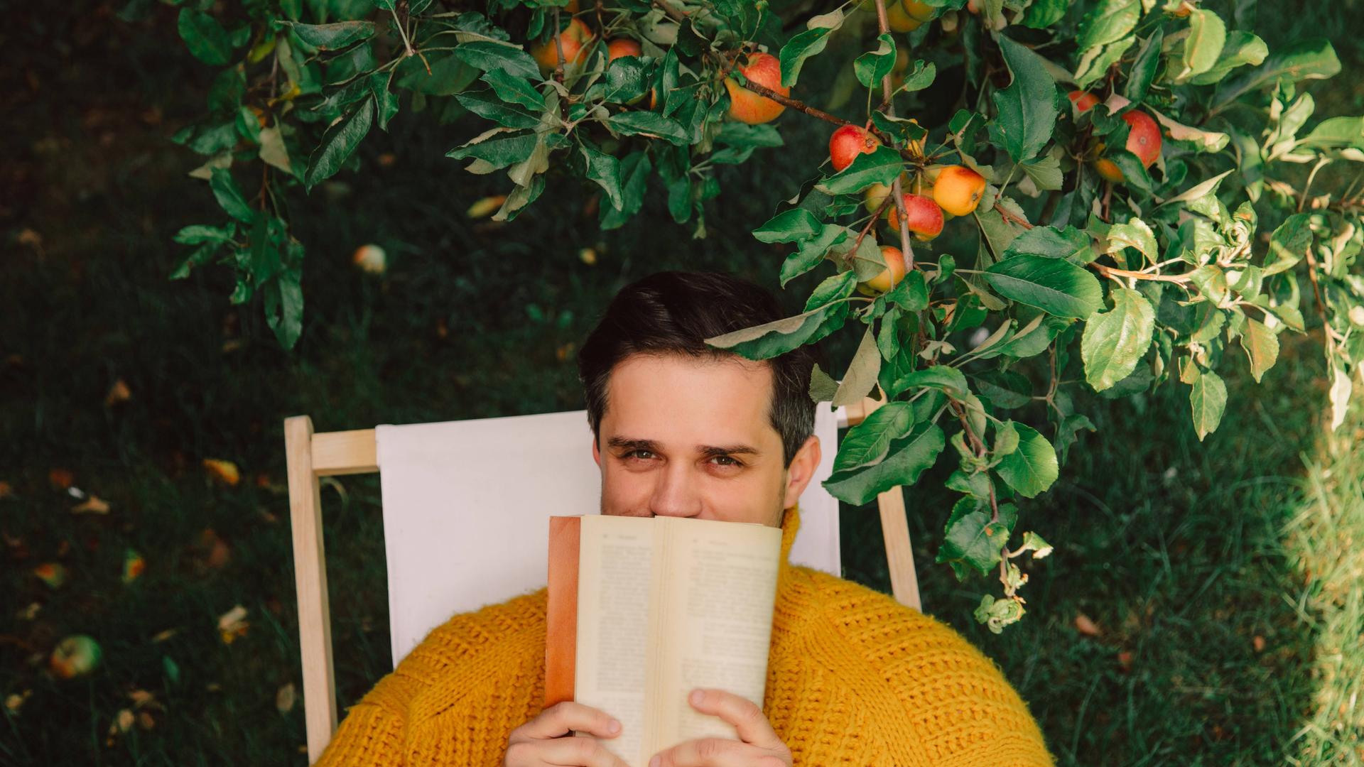 Ein Mann sitzt draußen in einem Liegestuhl unter einem Apfelbaum. Er hält sich ein Buch vor den Mund. Die Hälfte seines Gesichts ist dadruch verdeckt.