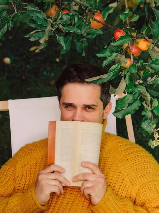 Ein Mann sitzt draußen in einem Liegestuhl unter einem Apfelbaum. Er hält sich ein Buch vor den Mund. Die Hälfte seines Gesichts ist dadruch verdeckt.