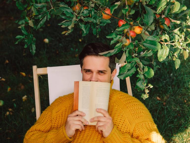 Ein Mann sitzt draußen in einem Liegestuhl unter einem Apfelbaum. Er hält sich ein Buch vor den Mund. Die Hälfte seines Gesichts ist dadruch verdeckt.
