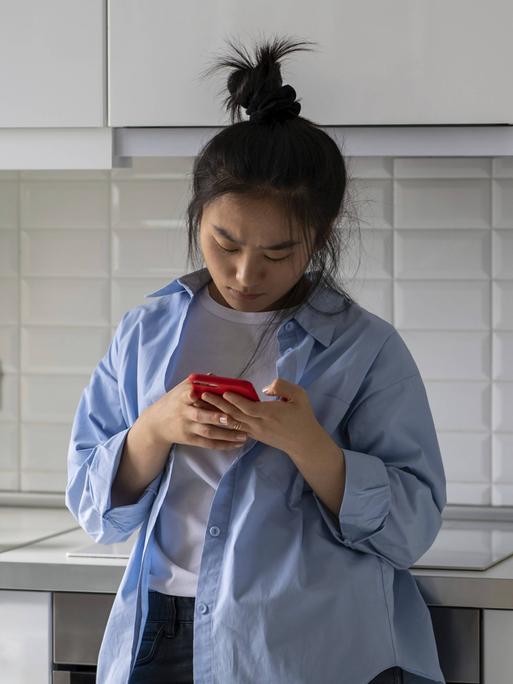 Eine junge Frau steht in einer Küche. Sie lehnt an der Küchenzeile an und schaut auf ihr Handy. Sie wirkt verärgert. Eine junge Frau steht in einer Küche. Sie lehnt an der Küchenzeile an und schaut auf ihr Handy. Sie wirkt verärgert.