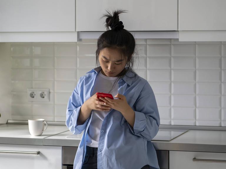 Eine junge Frau steht in einer Küche. Sie lehnt an der Küchenzeile an und schaut auf ihr Handy. Sie wirkt verärgert. Eine junge Frau steht in einer Küche. Sie lehnt an der Küchenzeile an und schaut auf ihr Handy. Sie wirkt verärgert.