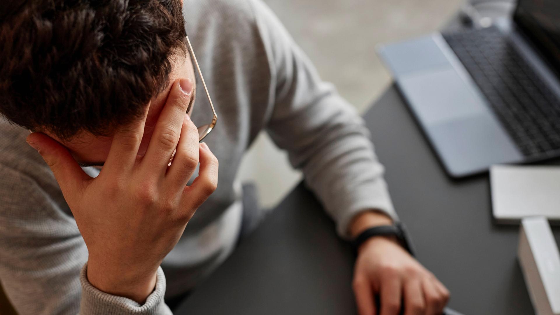Ein Mann sitzt vor einem Computer und einem Laptop an einem Schreibtisch vor. Wir sehen ihn von oben. Er hält seine Hand gegen seine Stirn.