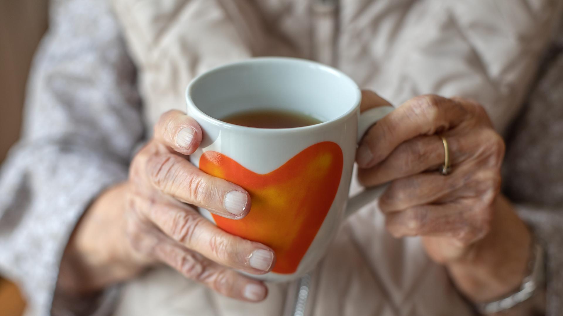 Die Hände einer älteren Frau halten eine Teetasse mit Herzmotiv.