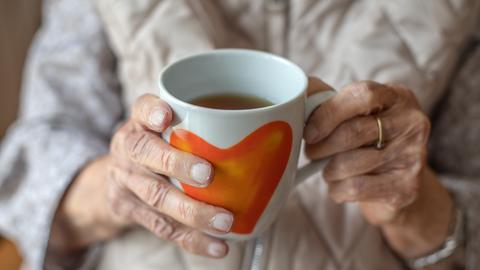 Die Hände einer älteren Frau halten eine Teetasse mit Herzmotiv.