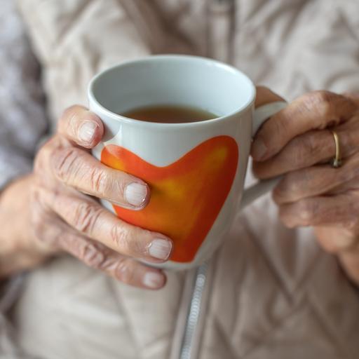 Die Hände einer älteren Frau halten eine Teetasse mit Herzmotiv.