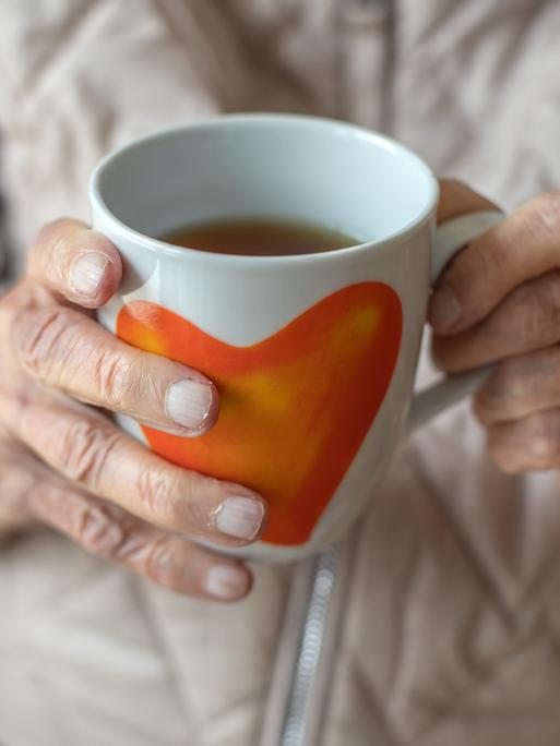 Die Hände einer älteren Frau halten eine Teetasse mit Herzmotiv.