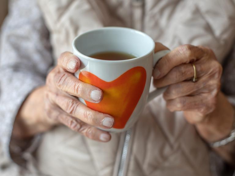 Die Hände einer älteren Frau halten eine Teetasse mit Herzmotiv.