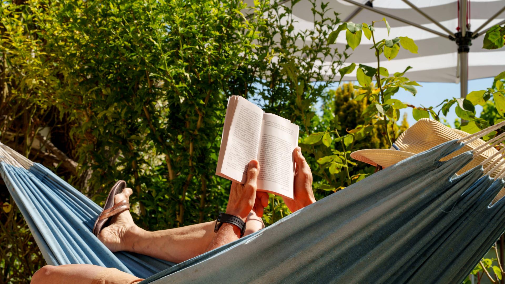 Eine Person mit Sonnenhut liegt in einer Hängematte und liest ein Buch. Eine Person mit Sonnenhut liegt in einer Hängematte und liest ein Buch.