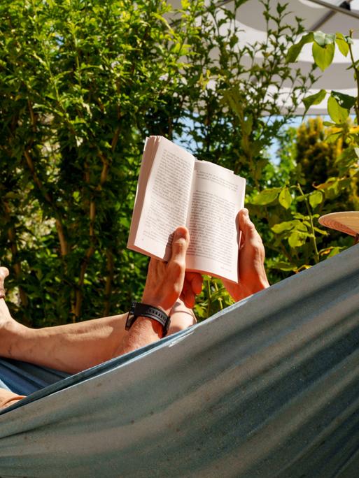 Eine Person mit Sonnenhut liegt in einer Hängematte und liest ein Buch.