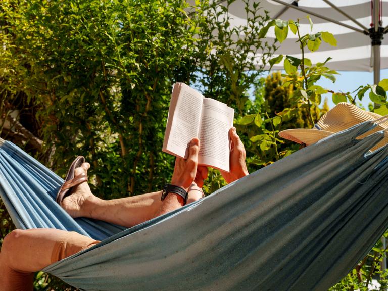 Eine Person mit Sonnenhut liegt in einer Hängematte und liest ein Buch.