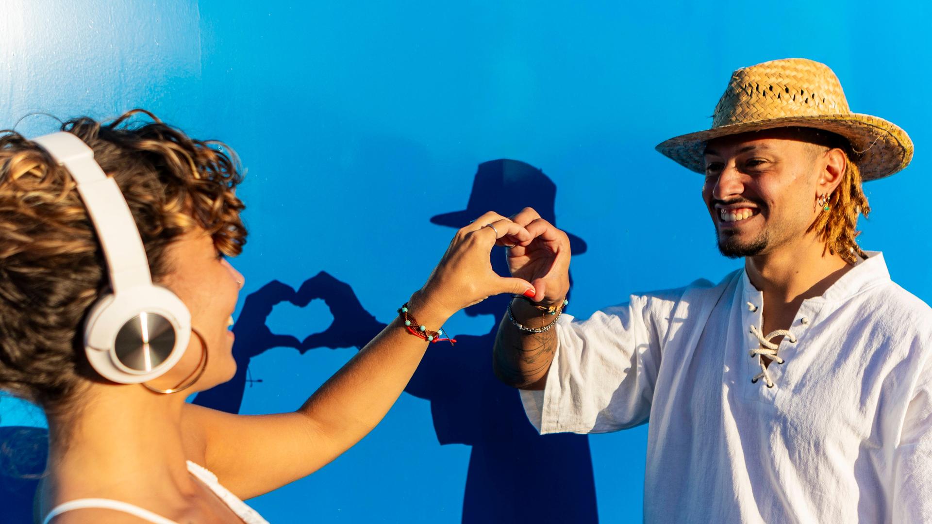 Ein Mann und eine Frau formen gemeinsam ein Herz mit ihren Händen vor einem blauen Hintergrund, die Frau trägt Kopfhörer Ein Mann und eine Frau formen gemeinsam ein Herz mit ihren Händen vor einem blauen Hintergrund, die Frau trägt Kopfhörer