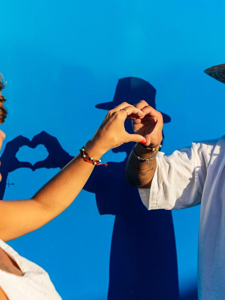 Ein Mann und eine Frau formen gemeinsam ein Herz mit ihren Händen vor einem blauen Hintergrund, die Frau trägt Kopfhörer