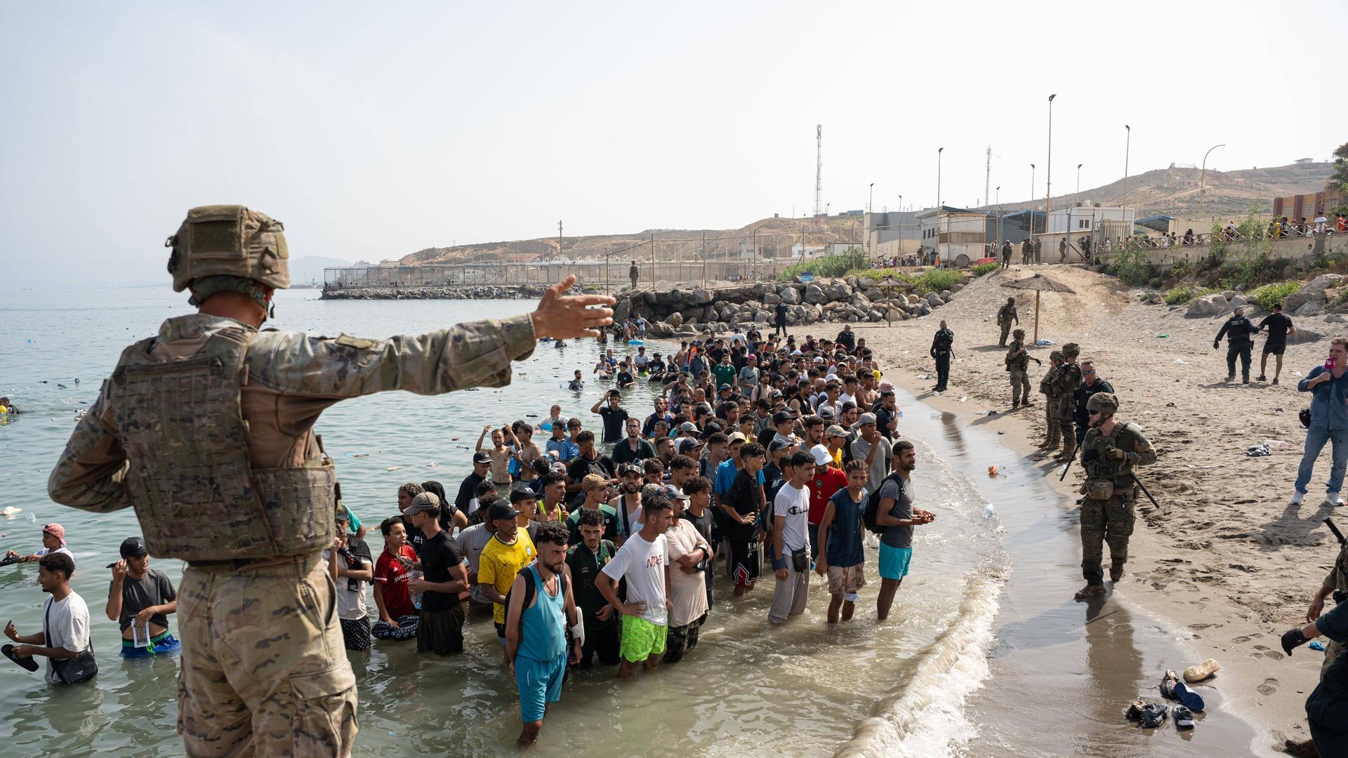 Geflüchtete stehen am Strand von Ceuta. Sie sind über das Meer von der marokkanischen Küste zu der spanischen Enklave geschwommen. Ein Soldat zeigt ihnen den Weg.