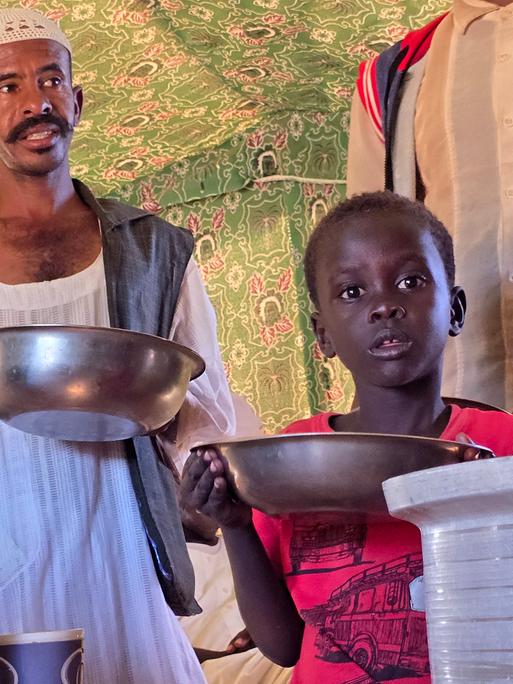 Ein Mann und ein Kind stehen mit Schüsseln in der Hand an einer Essensausgabe (Meroë/Sudan, 09.01.2025)
