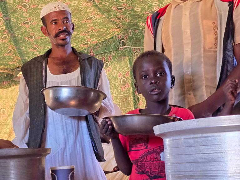 Ein Mann und ein Kind stehen mit Schüsseln in der Hand an einer Essensausgabe (Meroë/Sudan, 09.01.2025)