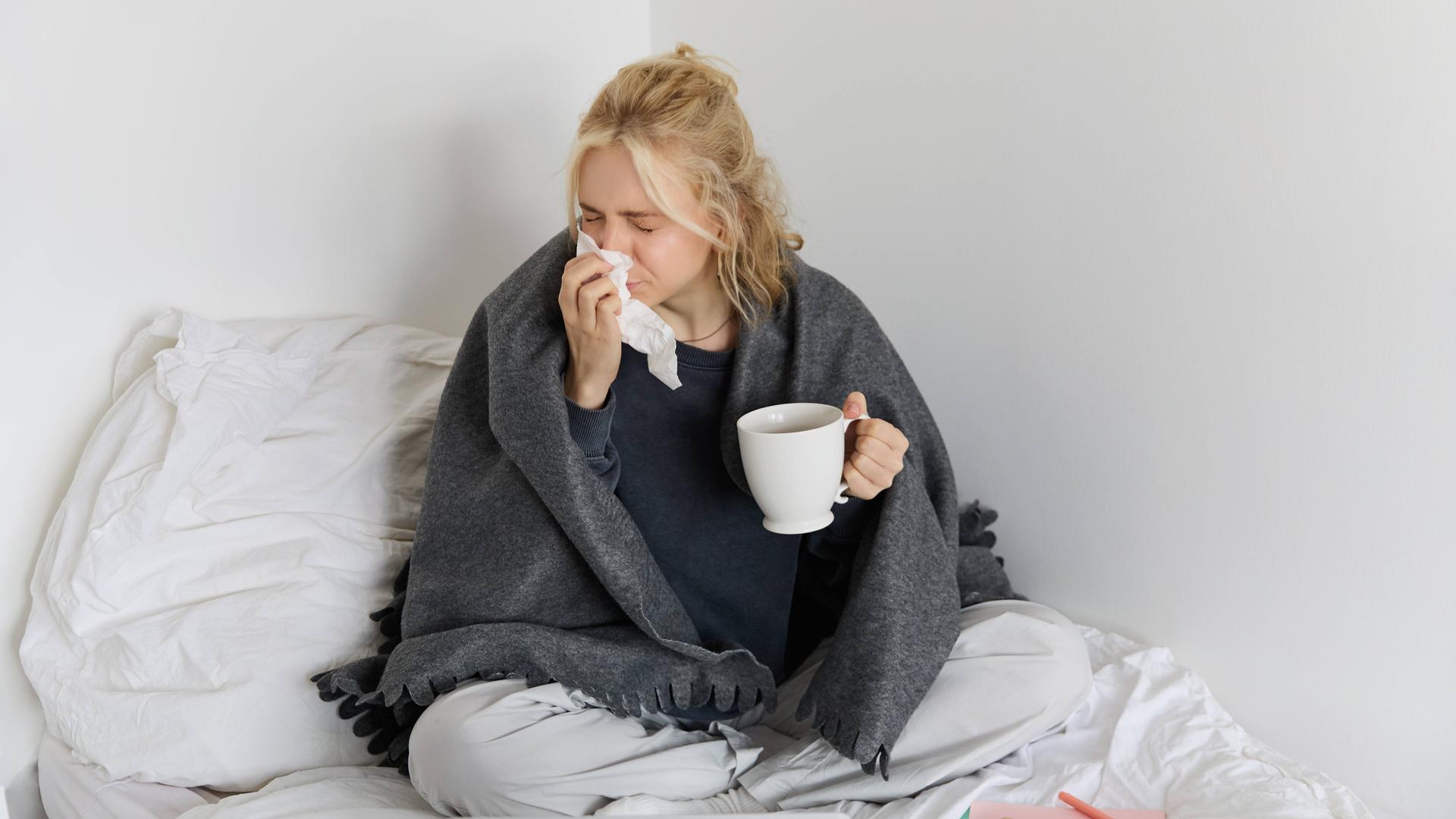 Eine Frau sitzt mit einer Decke um ihre Schultern auf einem Bett. Sie putzt sich mit einem Taschentuch die Nase und hält dabei eine Tasse in der Hand. Vor ihr auf dem Bett liegen ein Laptop und Notizbücher. Eine Frau sitzt mit einer Decke um ihre Schultern auf einem Bett. Sie putzt sich mit einem Taschentuch die Nase und hält dabei eine Tasse in der Hand. Vor ihr auf dem Bett liegen ein Laptop und Notizbücher.