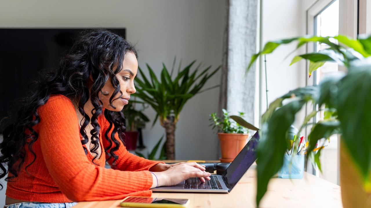 Eine junge Frau in einem orangefarbenen Pulli sitzt an einem Holztisch und arbeitet an einem Laptop. Ein Handy liegt neben dem Computer auf dem Tisch.