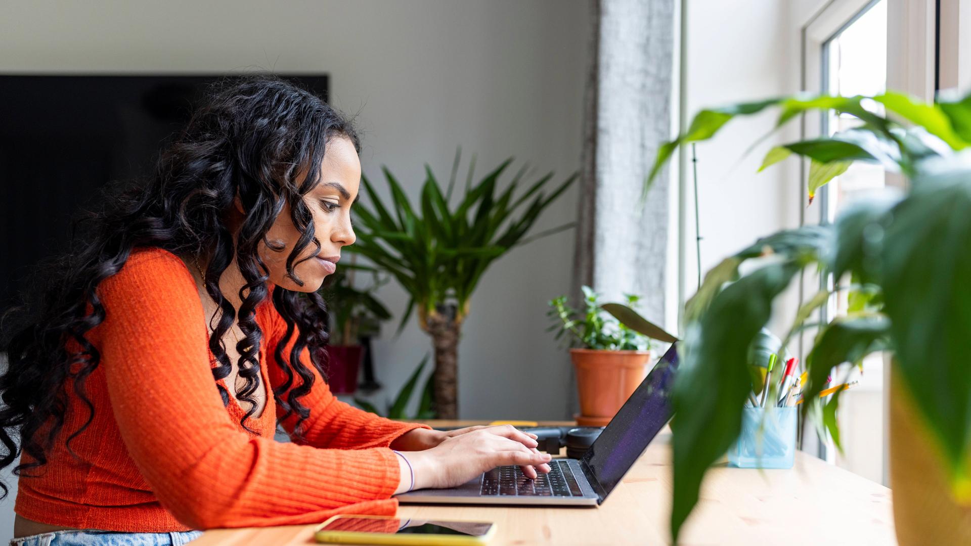 Eine junge Frau in einem orangefarbenen Pulli sitzt an einem Holztisch und arbeitet an einem Laptop. Ein Handy liegt neben dem Computer auf dem Tisch. Eine junge Frau in einem orangefarbenen Pulli sitzt an einem Holztisch und arbeitet an einem Laptop. Ein Handy liegt neben dem Computer auf dem Tisch.