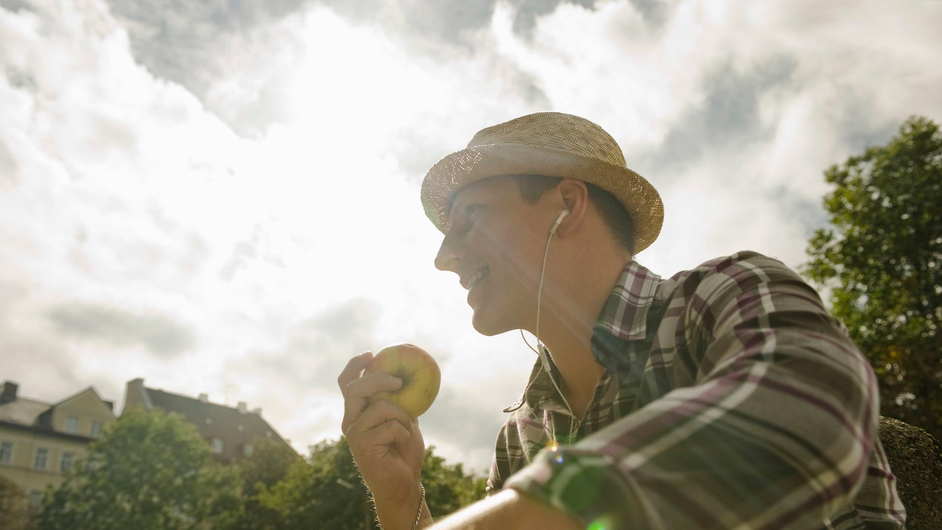 Ein Mann sitzt in der Sonne, trägt einen Strohhut und hält einen Apfel in der Hand (Symbolfoto) Ein Mann sitzt in der Sonne, trägt einen Strohhut und hält einen Apfel in der Hand (Symbolfoto)