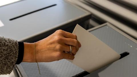 Gezeigt ist eine Hand, die einen Stimmzettel in eine Wahlurne wirft.