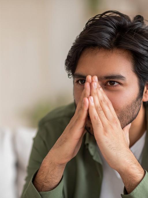 Ein Mann sitzt auf einer Couch und hält sich seine Hände vor das Gesicht, dadurch wird seine Nase verdeckt. Ein Mann sitzt auf einer Couch und hält sich seine Hände vor das Gesicht, dadurch wird seine Nase verdeckt.