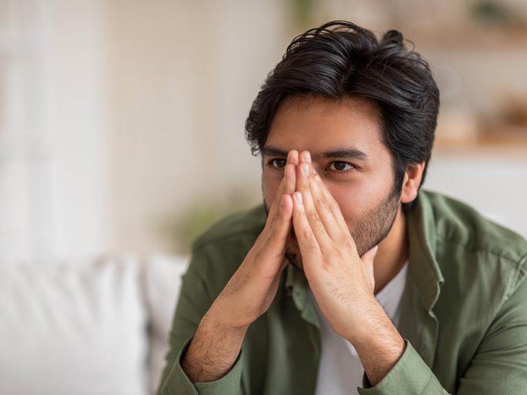 Ein Mann sitzt auf einer Couch und hält sich seine Hände vor das Gesicht, dadurch wird seine Nase verdeckt. Ein Mann sitzt auf einer Couch und hält sich seine Hände vor das Gesicht, dadurch wird seine Nase verdeckt.