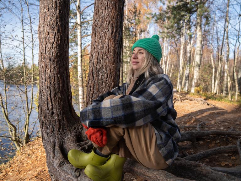 Middle aged woman contemplating enjoying nature in park sitting on tree roots. Relaxed female in warm clothes resting in autumn forest on lake shore. Slow life, retreat concept Middle aged woman contemplating enjoying nature in park sitting on tree roots. Relaxed female in warm clothes resting in autumn forest on lake shore. Slow life, retreat concept