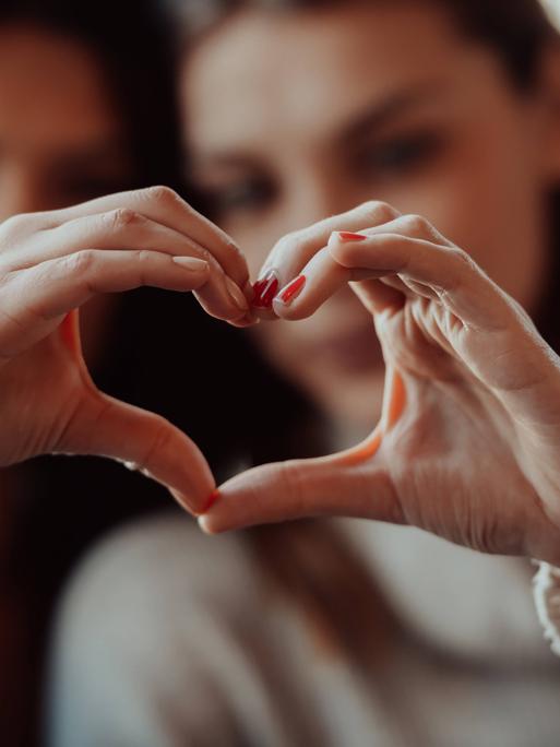 Zwei Frauen halten ihre Hände so zusammen, dass sie gemeinsam ein Herz ergeben. Die Herz-Hand ist im Vordergrund. Die beiden Frauen sind unscharf im Hintergrund.