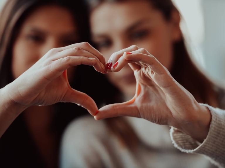 Zwei Frauen halten ihre Hände so zusammen, dass sie gemeinsam ein Herz ergeben. Die Herz-Hand ist im Vordergrund. Die beiden Frauen sind unscharf im Hintergrund.