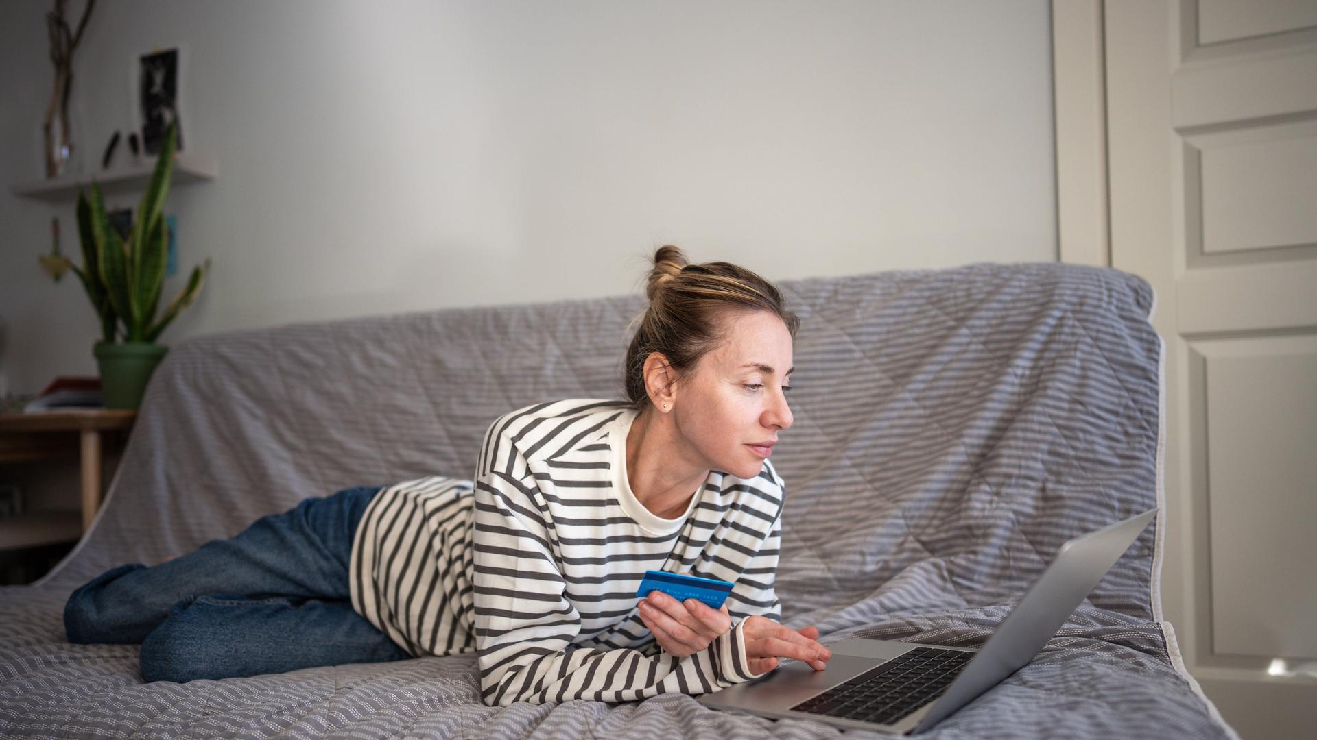 Eine Frau, mit einer Bankkarte in der Hand, liegt auf der Couch und benutzt einen Laptop
