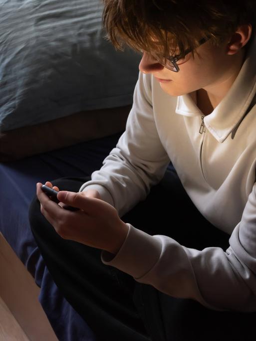 Ein junger Mann sitzt auf seinem Bett und schaut in sein Smartphone