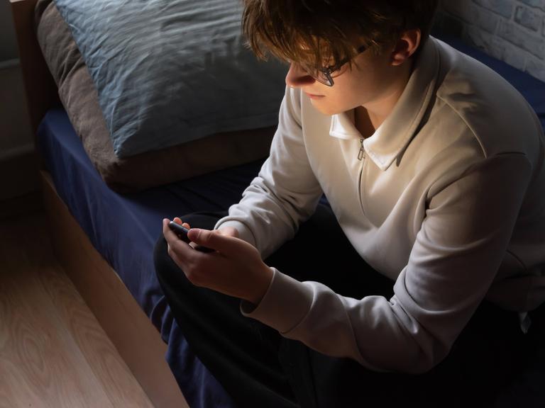 Ein junger Mann sitzt auf seinem Bett und schaut in sein Smartphone