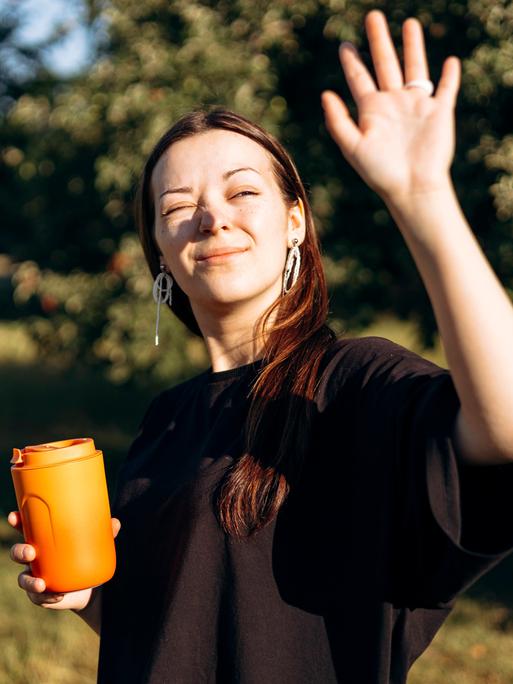 Eine Frau ist an einem sonnigen Tag draußen unterwegs. Sie hält einen orangefarbenen Thermobecher in der einen Hand und die andere Hand vor ihr Gesicht, um sich vor der Sonne zu schützen.