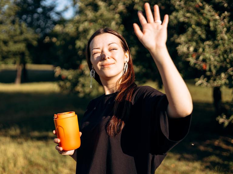Eine Frau ist an einem sonnigen Tag draußen unterwegs. Sie hält einen orangefarbenen Thermobecher in der einen Hand und die andere Hand vor ihr Gesicht, um sich vor der Sonne zu schützen.
