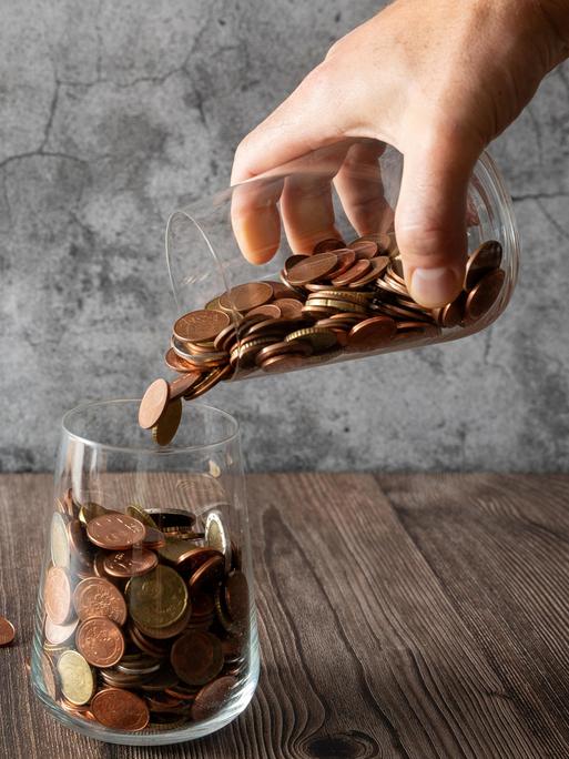 Eine Hand schüttet Münzen aus einem Glas in ein anderes