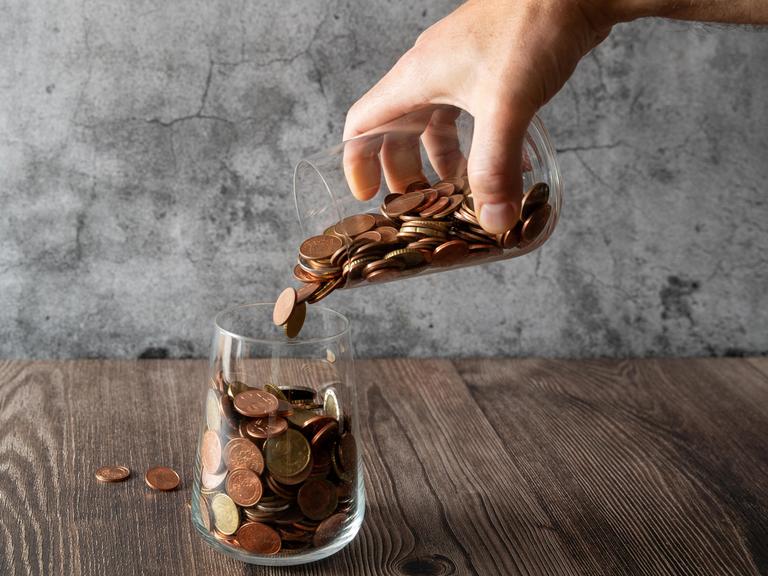 Eine Hand schüttet Münzen aus einem Glas in ein anderes