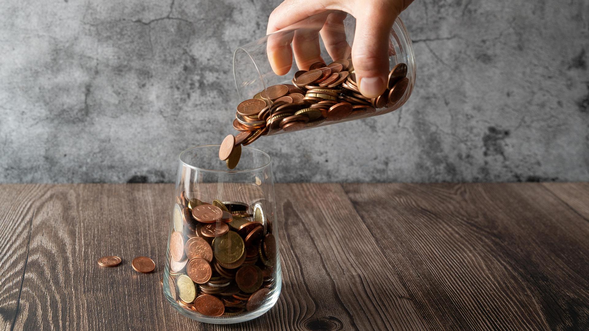 Hand schüttet Münzen aus einem Glas in ein anderes Hand schüttet Münzen aus einem Glas in ein anderes