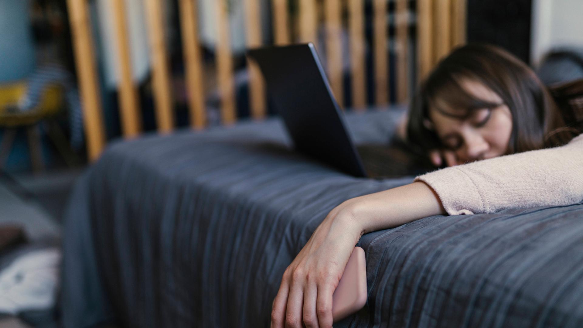Eine Frau liegt auf dem Bett, die Hand, in der sie ein Smartphone hält, hängt schlaff herab, Vor ihr ein Laptop.