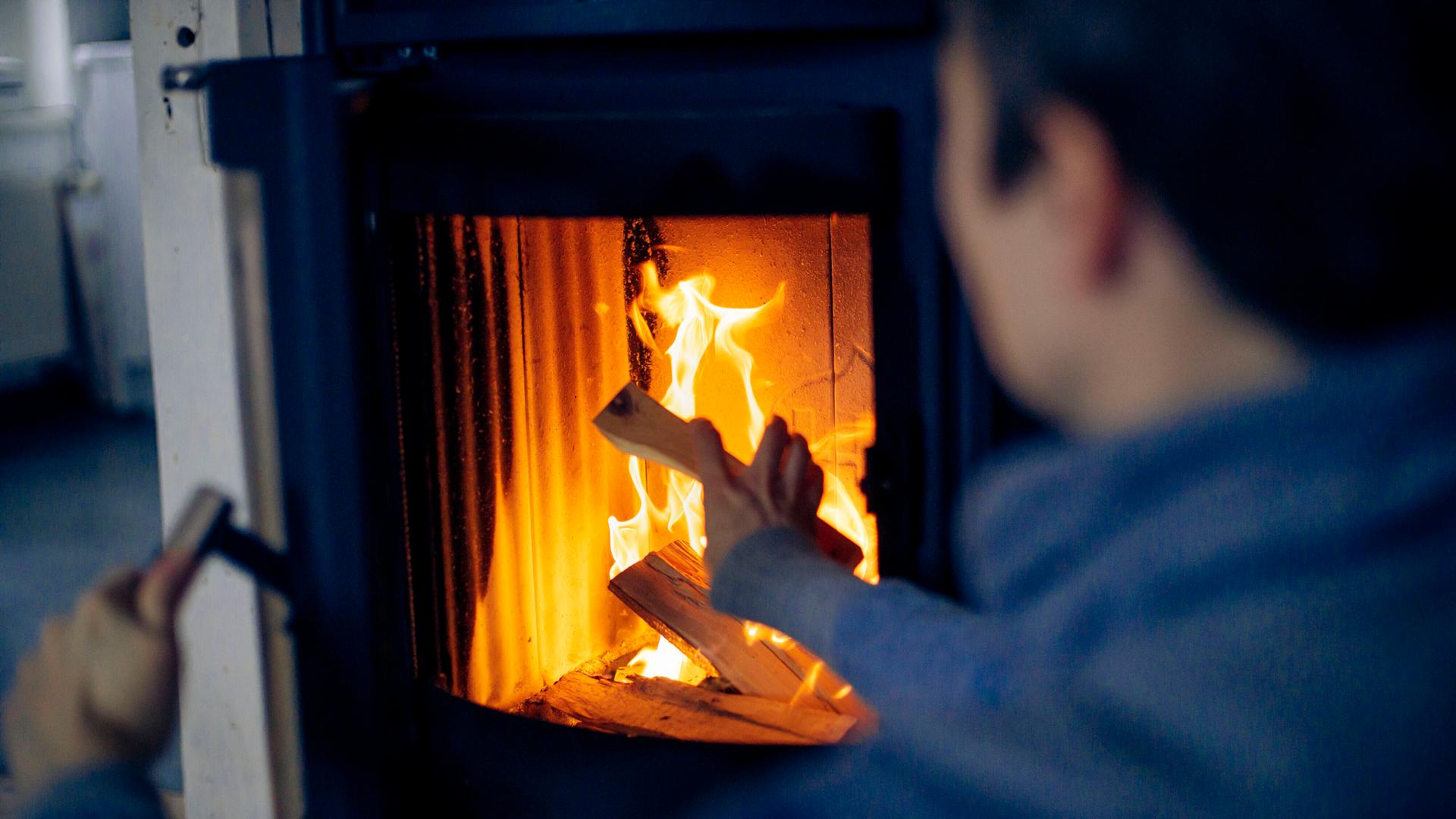Eine Person sitzt vor einem Kaminofen, darin brennendes Holz. Der Mann hat die Kamintür geöffnet und legt Holz nach.