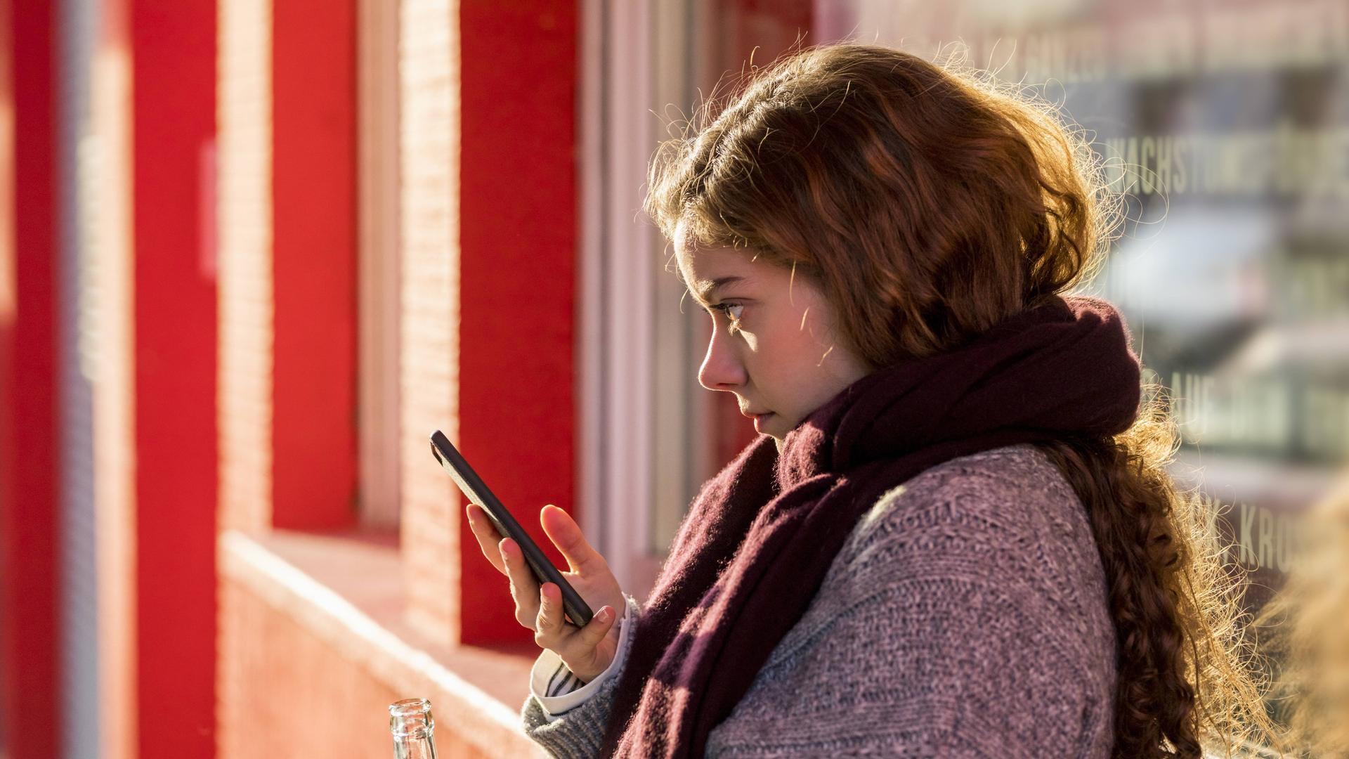 Eine Jugendliche steht auf einer roten Brücke. Sie hat in der einen Hand ein Handy, in der anderen eine Limonade. Eine Jugendliche steht auf einer roten Brücke. Sie hat in der einen Hand ein Handy, in der anderen eine Limonade.