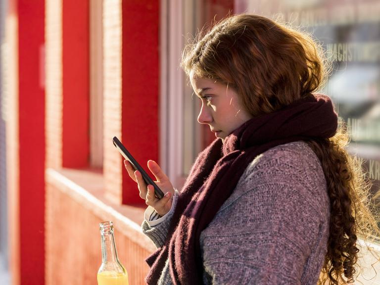 Eine Jugendliche steht auf einer roten Brücke. Sie hat in der einen Hand ein Handy, in der anderen eine Limonade.