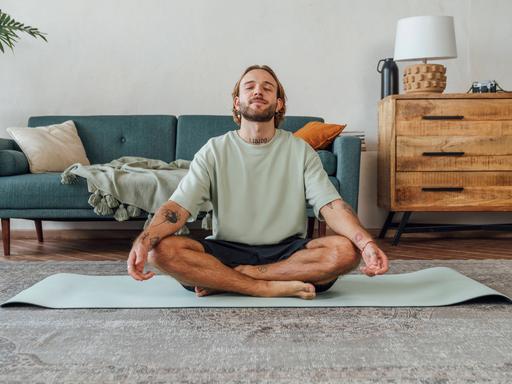 Ein Mann sitzt zu Hause im Schneidersitz auf dem Boden und meditiert Ein Mann sitzt zu Hause im Schneidersitz auf dem Boden und meditiert