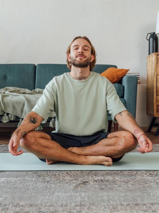 Ein Mann sitzt zu Hause im Schneidersitz auf dem Boden und meditiert Ein Mann sitzt zu Hause im Schneidersitz auf dem Boden und meditiert
